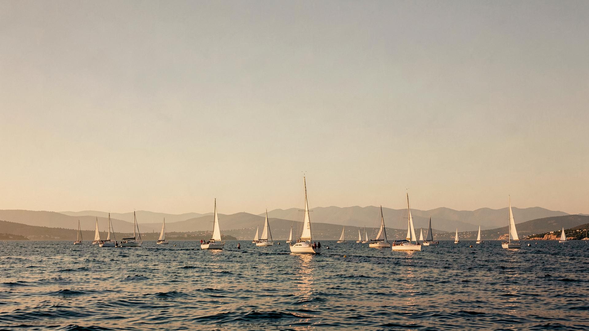 A racing fleet crossing a coastal course under open sky, land visible on the horizon.